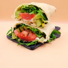 Dietetyczna tortilla z warzywami: sałatą, pomidorem, ogórkiem i rukolą, na ciemnej desce.